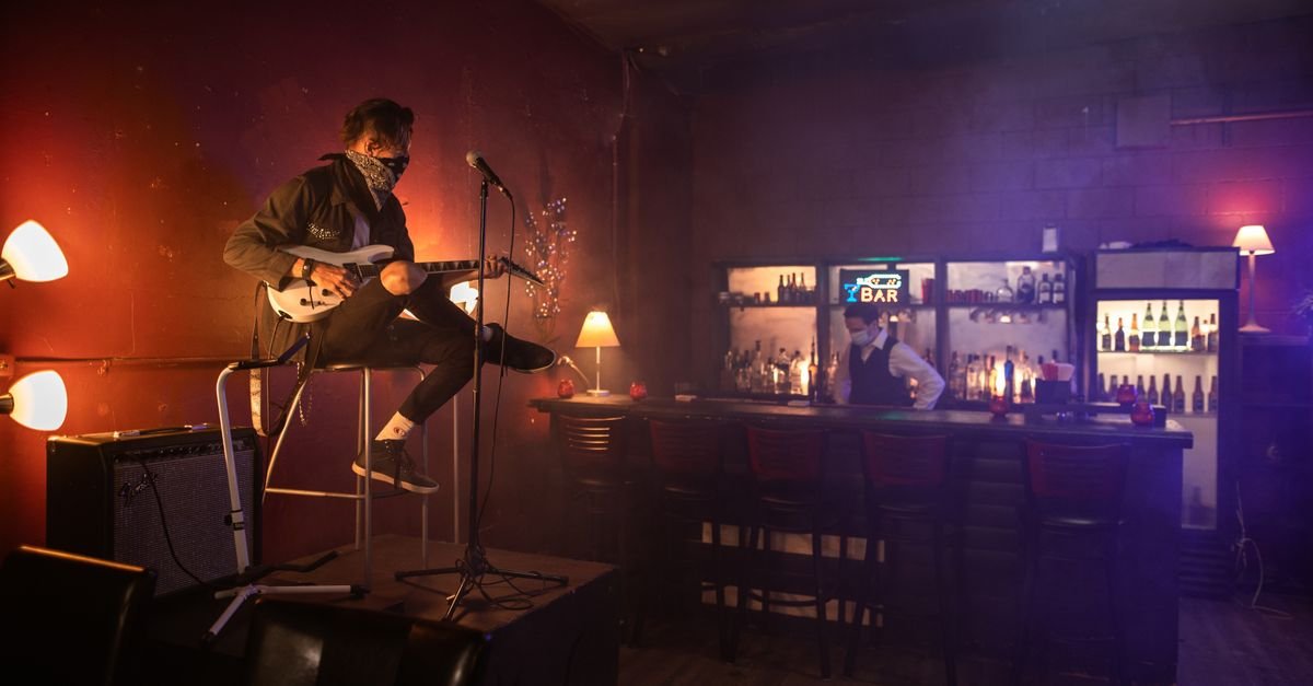 Cantor com guitarra se apresenta sentado em palco de bar com luz baixa