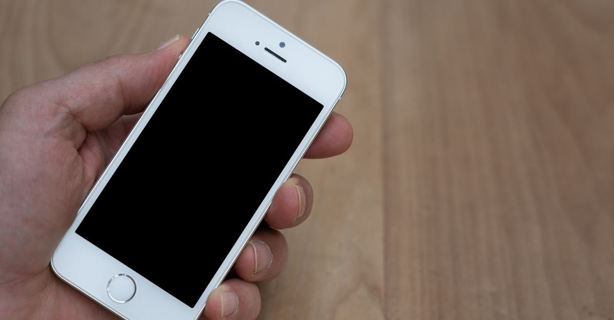 Mão segurando um smartphone com a tela desligada sobre fundo de madeira