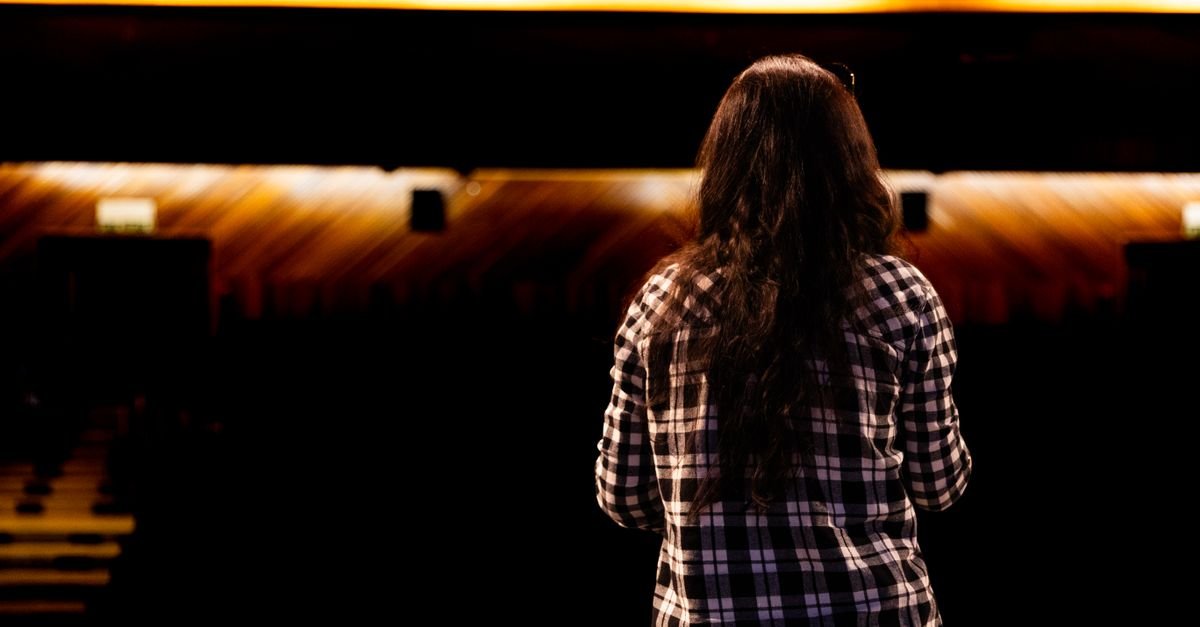 Pessoa de costas em pé no palco de um teatro vazio, com plateia ao fundo e luz quente