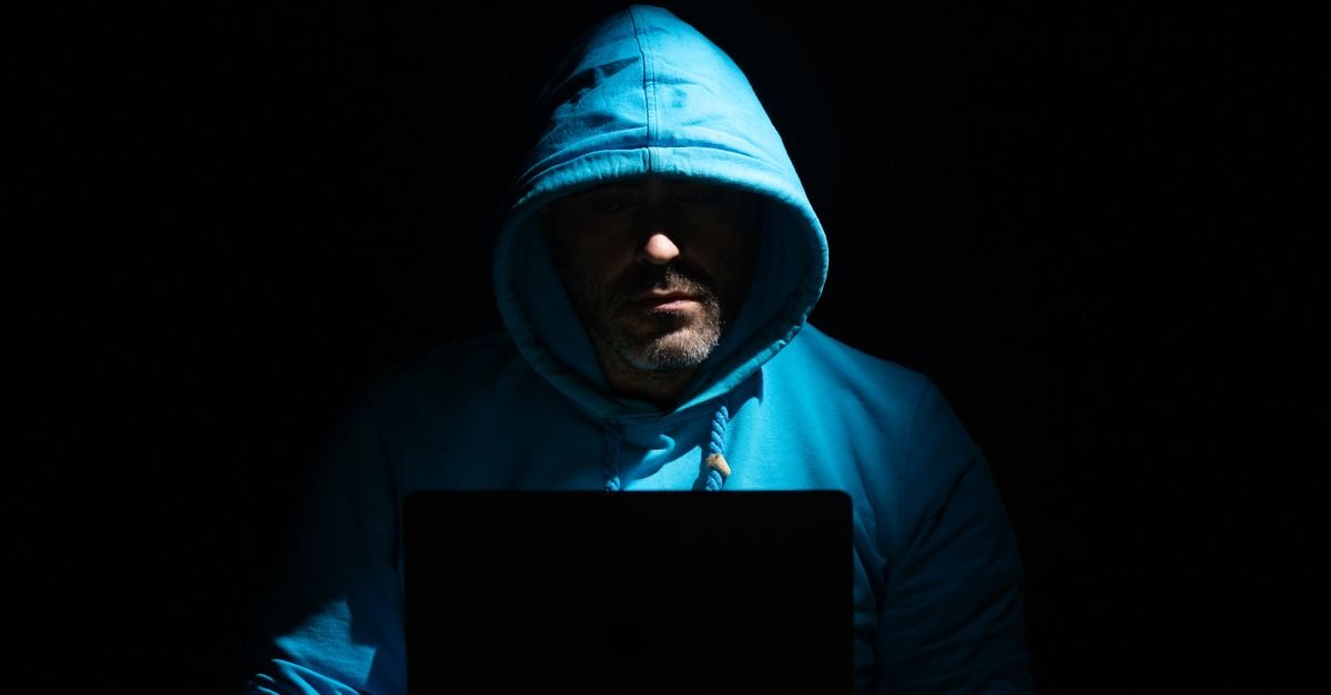 Homem de capuz diante de um notebook em ambiente escuro, ilustrando fraude digital