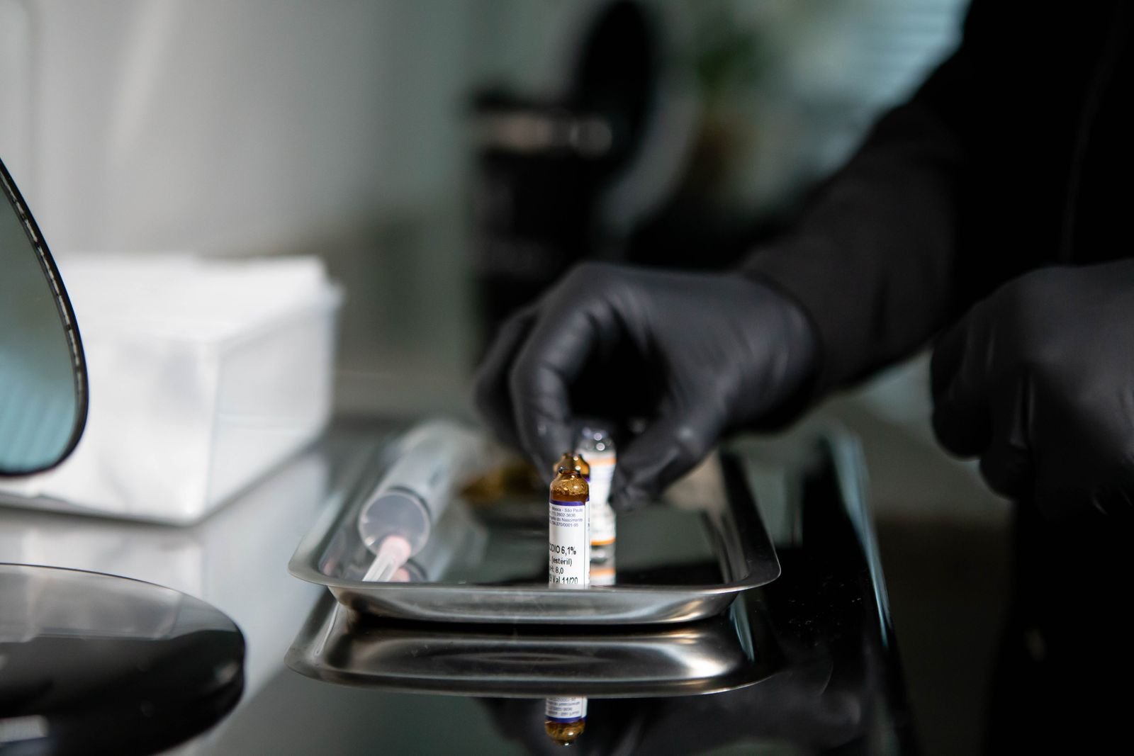 Mãos com luva preta manuseiam ampolas de medicamento e uma seringa sobre bandeja de inox