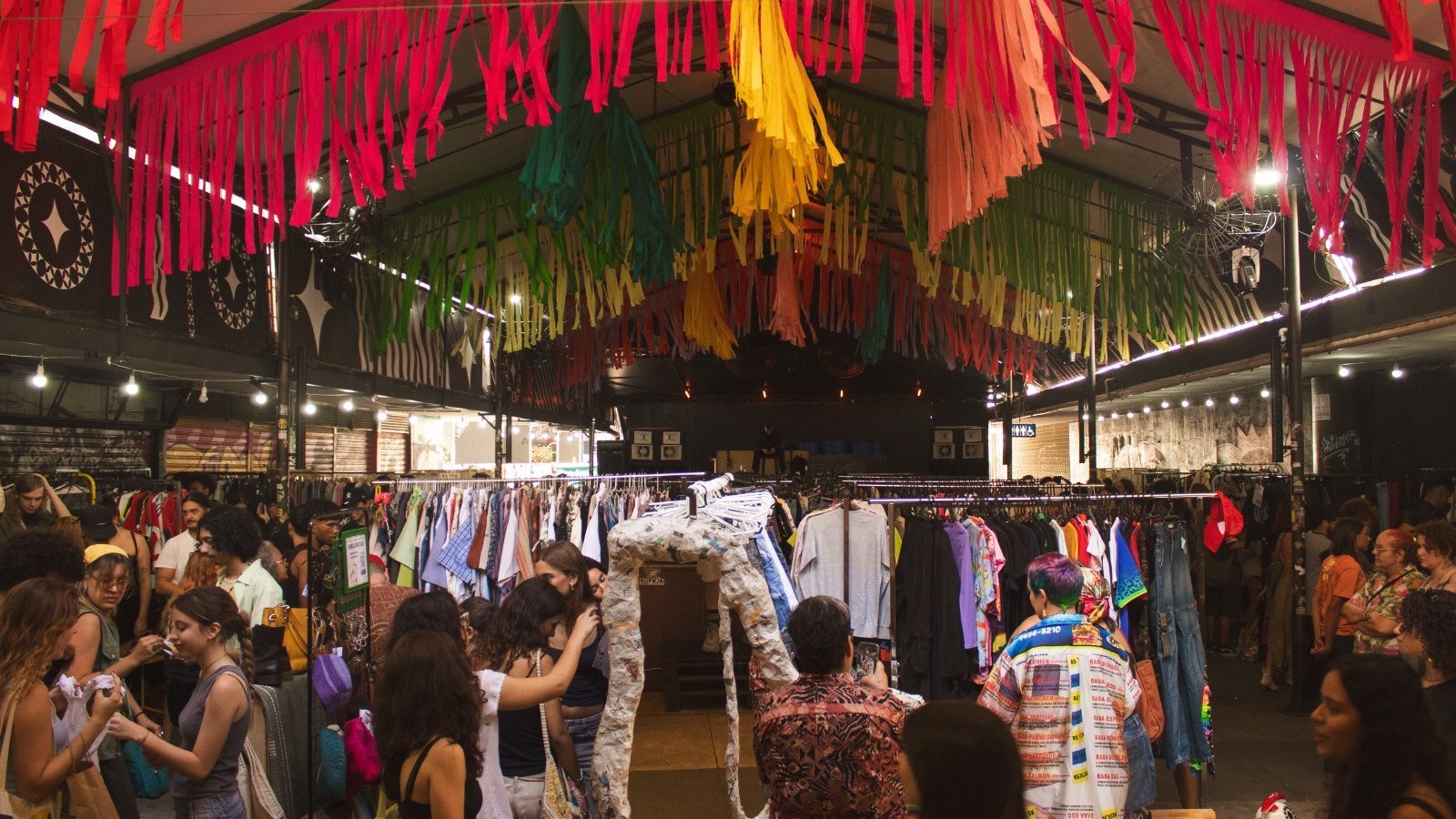 Brechó com araras de roupas e decoração de franjas coloridas, com público adulto circulando