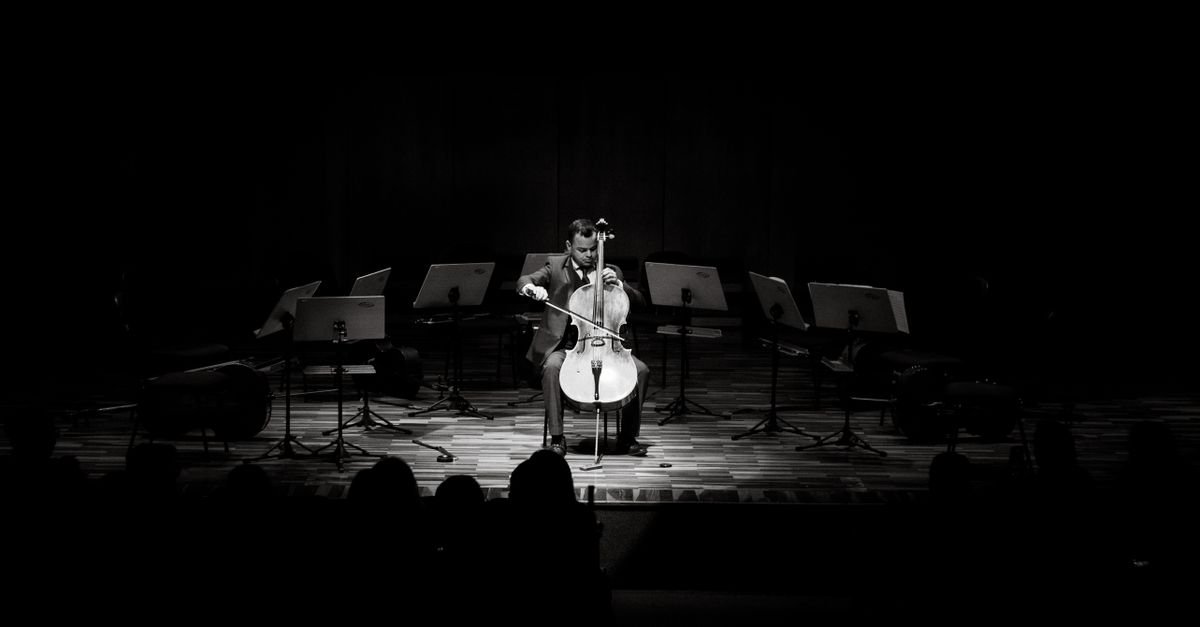 Violoncelista sozinho no palco iluminado, em foto em preto e branco, durante apresentação