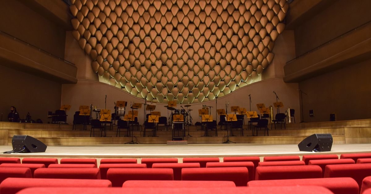Palco montado visto da plateia de poltronas vermelhas, com estantes de partitura e instrumentos
