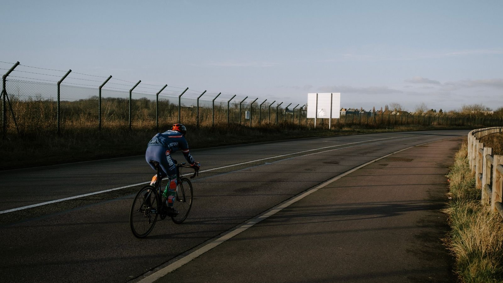 Ciclista pedala sozinho em pista de asfalto vazia, ao lado de cerca e mureta