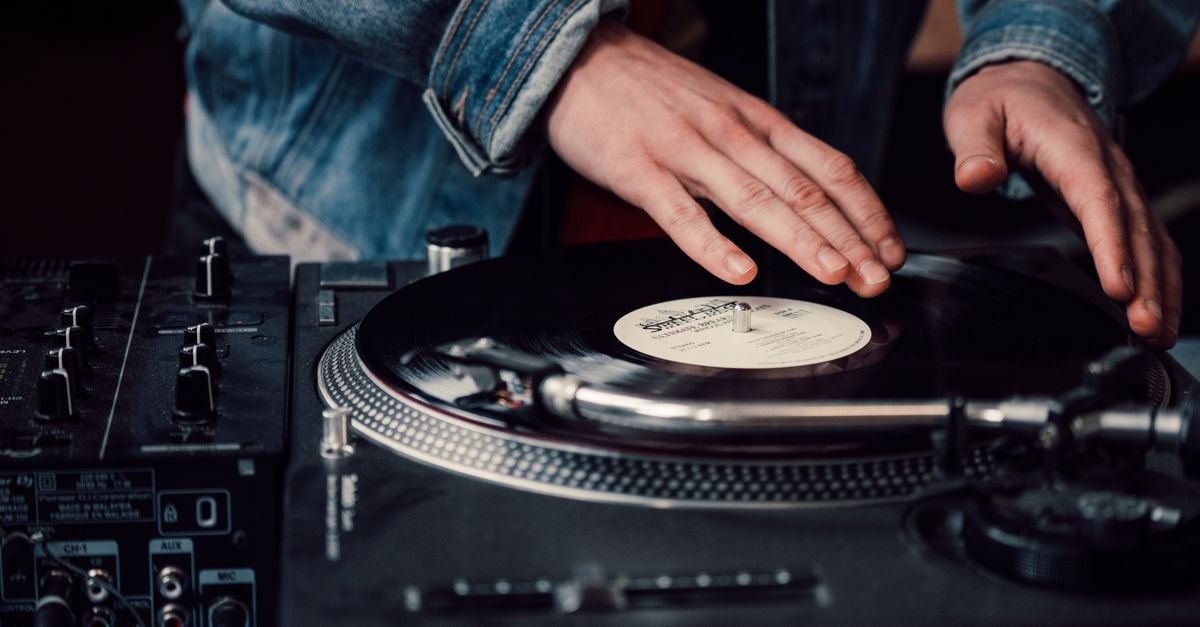 Mãos de DJ sobre disco de vinil em toca-discos, ao lado de mesa de mixagem