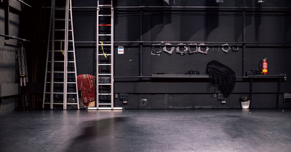 Bastidor de teatro vazio com escadas metálicas, refletores guardados e piso de palco escuro