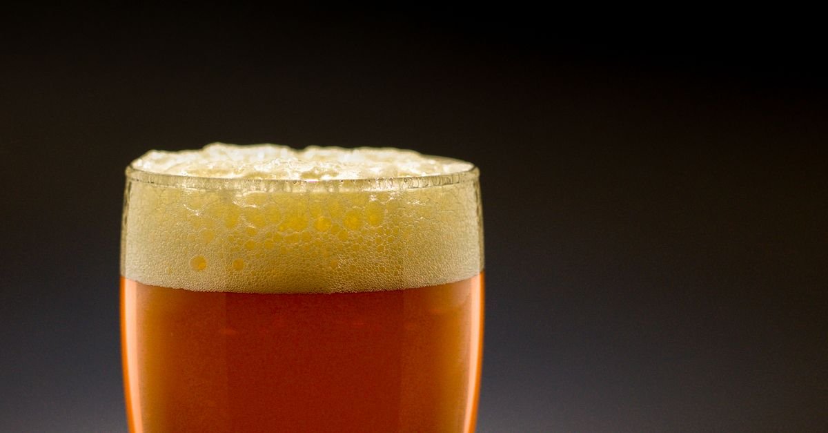 Copo de cerveja âmbar com colarinho de espuma em primeiro plano, sobre fundo escuro