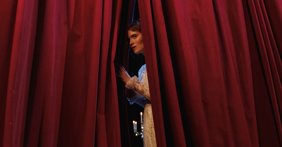 Atriz em traje de época observa a plateia entre cortinas de veludo vermelho de um teatro