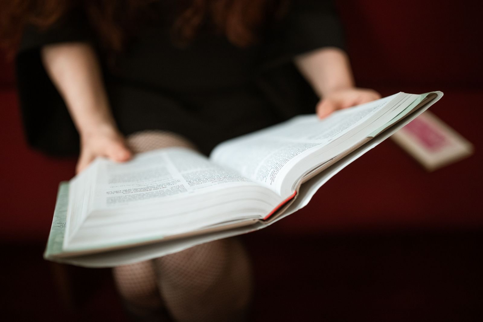 Mulher com um livro grosso aberto nas mãos, sentada em um sofá vermelho