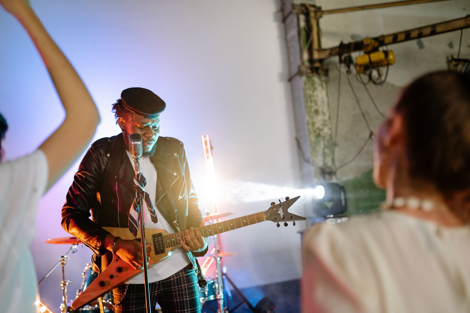 Guitarrista de jaqueta de couro toca guitarra em formato V diante de um microfone em show de rock, com plateia em primeiro plano