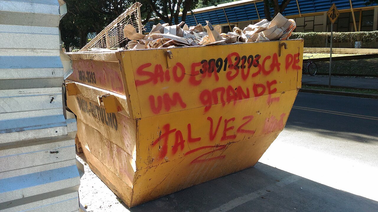 Caçamba amarela cheia de entulho estacionada em via pública com telefone da empresa pintado na lateral