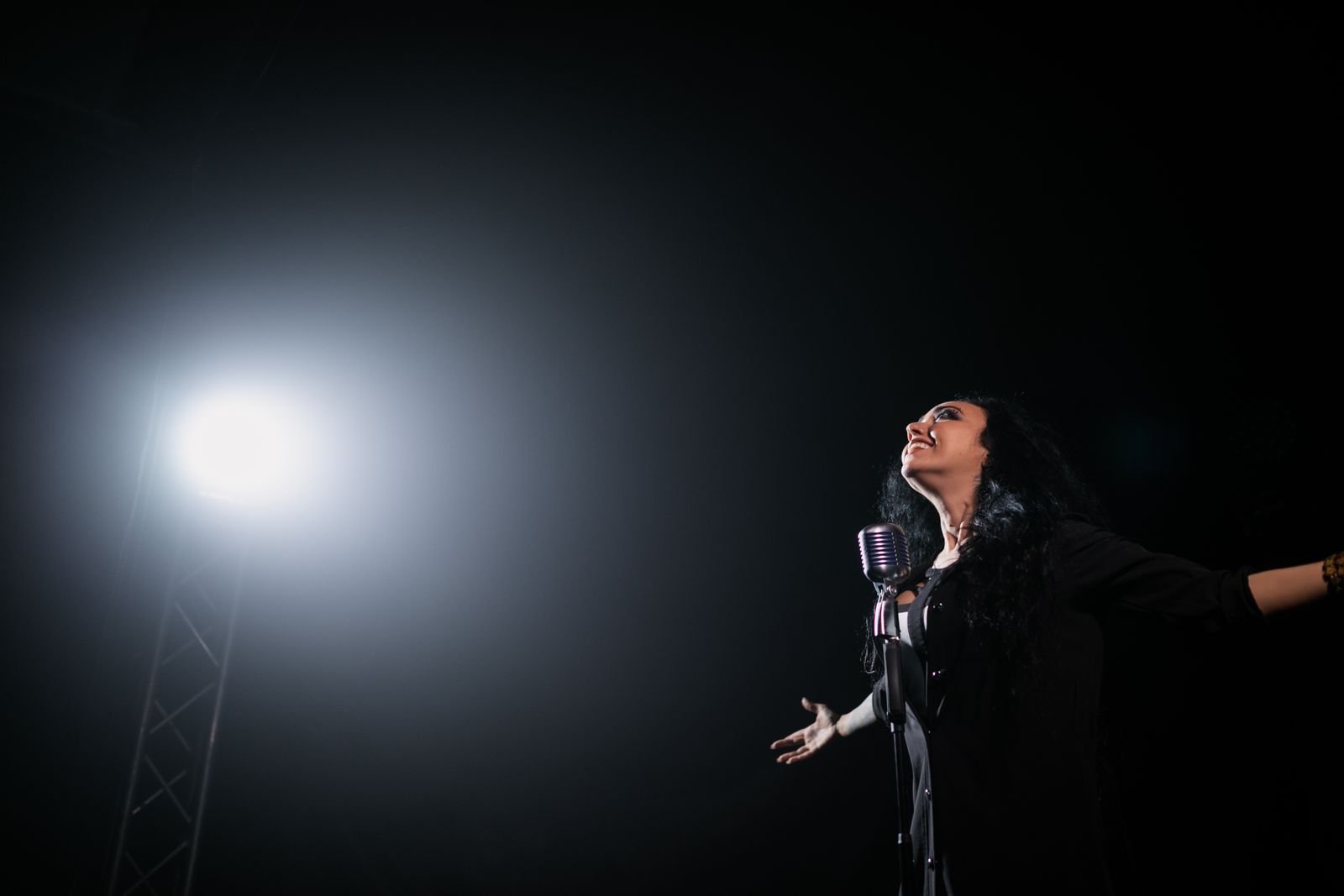 Cantora de braços abertos ao lado de um microfone antigo, sob um refletor, em palco escuro