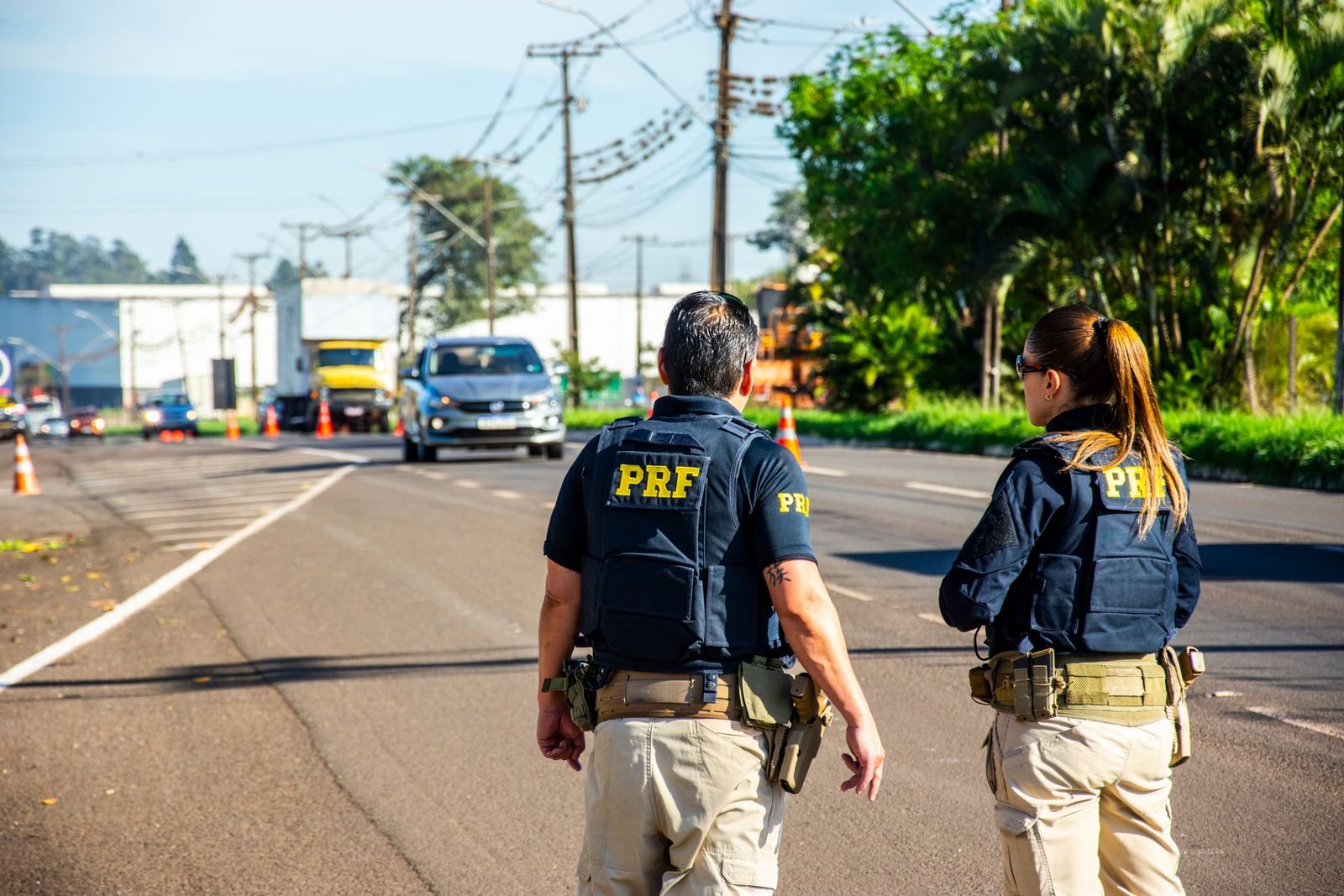Dois agentes da Polícia Rodoviária Federal de costas, em ponto de fiscalização com cones, enquanto carros e um caminhão se aproximam