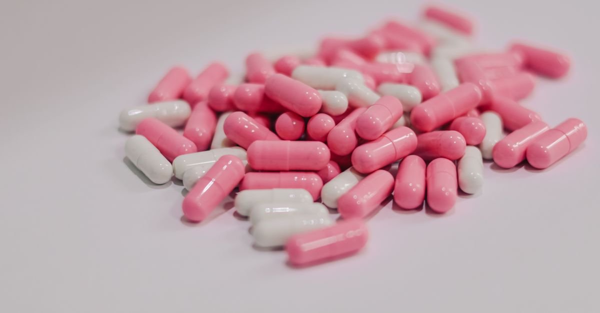 Cápsulas farmacêuticas rosas e brancas espalhadas sobre superfície branca, em close