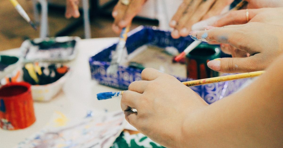 Mãos de participantes de uma oficina segurando pincéis sobre mesa com tintas e potes coloridos