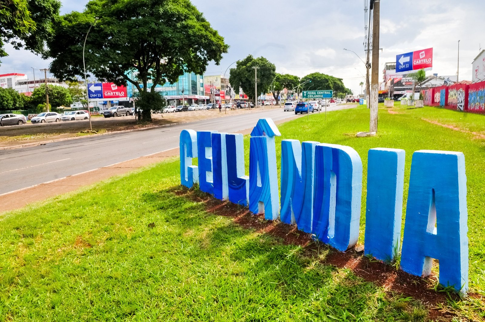 Letreiro azul com o nome Ceilândia em canteiro gramado, ao lado de avenida movimentada