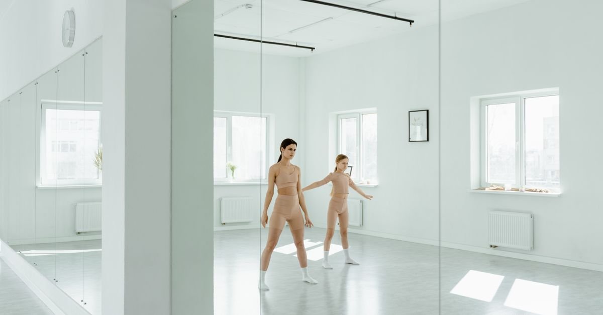 Duas bailarinas ensaiam em sala de dança com piso claro e parede de espelhos, em imagem ilustrativa