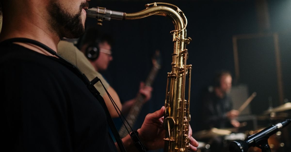 Saxofonista em primeiro plano tocando no palco, com baixista e baterista desfocados ao fundo