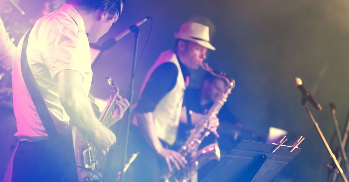 Baixista e saxofonista tocando em palco iluminado por luz roxa, com estante de partitura à frente