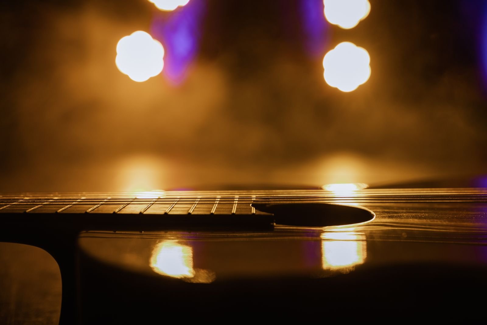 Violão acústico em close sobre um palco, com luzes douradas e roxas desfocadas ao fundo (foto ilustrativa)