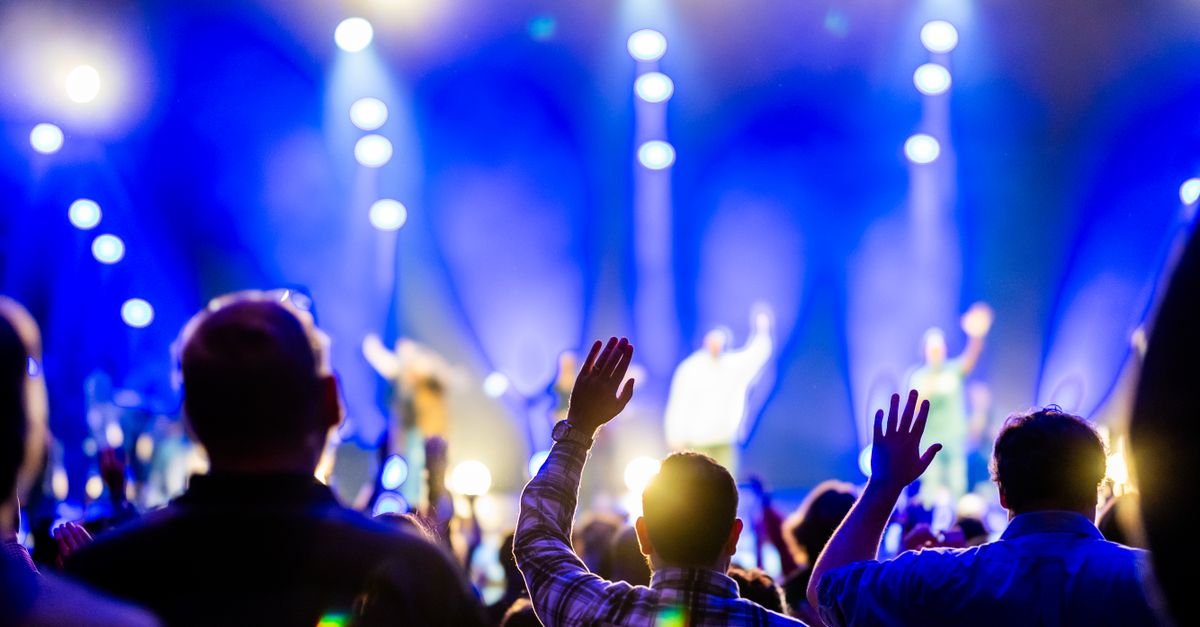 Público de costas com braços erguidos diante de palco iluminado em azul