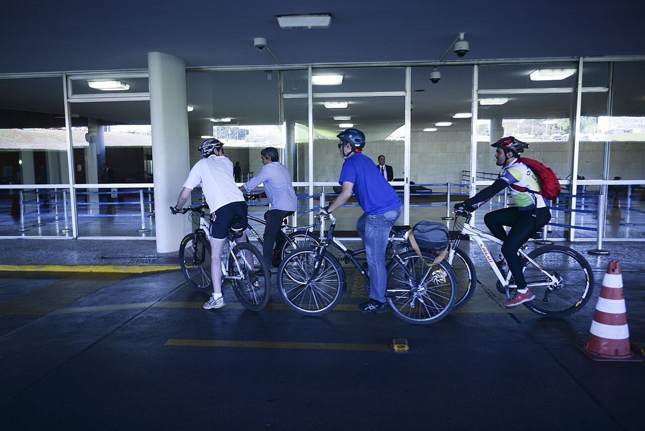 Três ciclistas com bicicletas e capacetes em uma entrada coberta de prédio público em Brasília