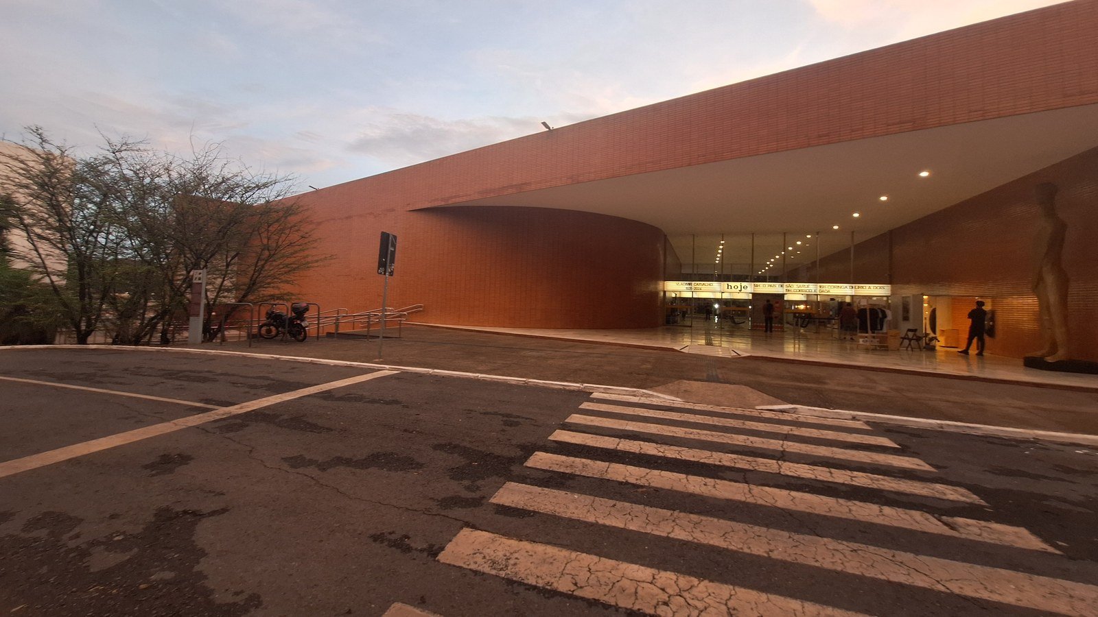 Fachada curva de tijolo do Cine Brasília ao anoitecer, com a marquise iluminada e público na entrada