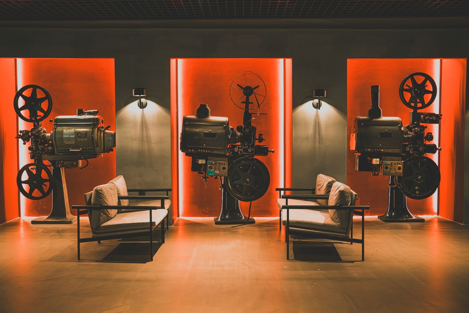 Três projetores de cinema antigos iluminados em vermelho ao lado de poltronas, em sala de exibição