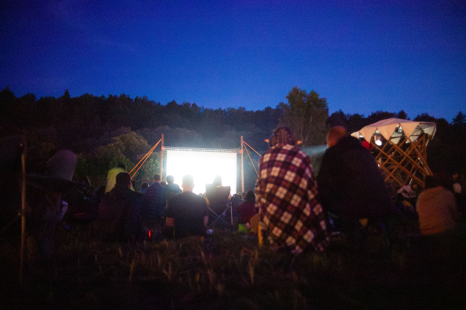 Público sentado na grama assiste a filme em tela ao ar livre à noite