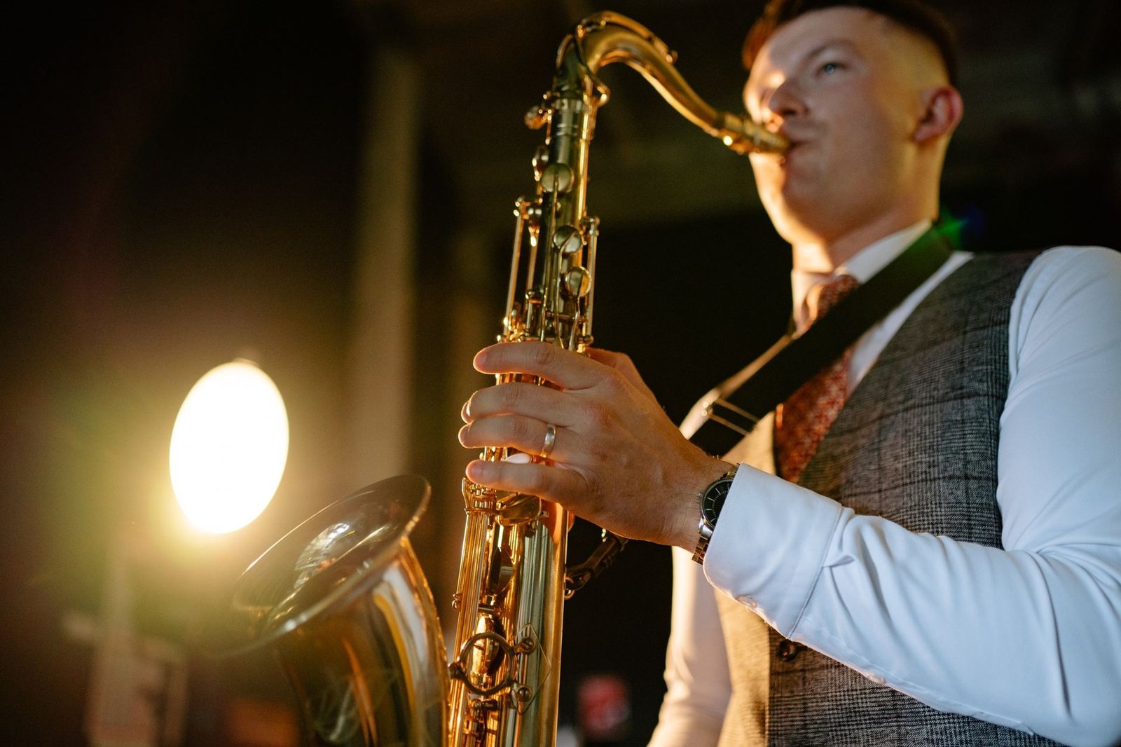 Saxofonista de colete toca saxofone tenor diante de um refletor aceso em palco escuro