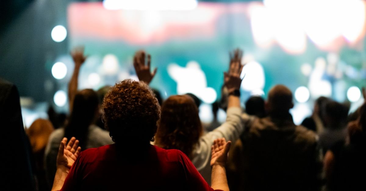 Publico de costas com as maos levantadas durante culto em evento fechado com luzes de palco