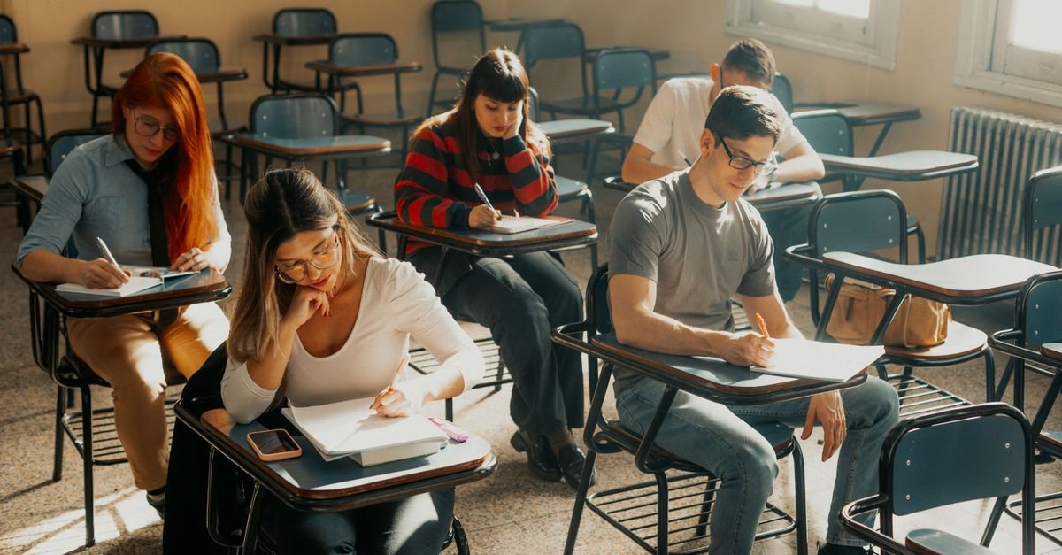 Candidatos sentados em carteiras de sala de aula escrevendo durante aplicação de prova