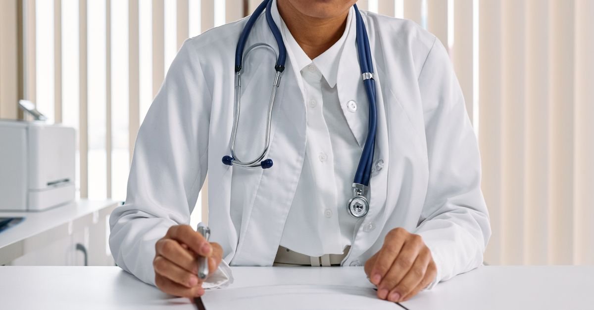 Profissional de saúde de jaleco branco com estetoscópio sentada à mesa de consultório