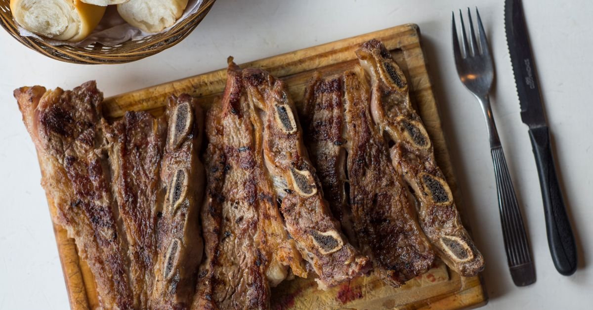 Costela bovina grelhada servida em tábua de madeira com pão, no estilo do asado argentino