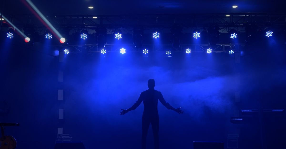 Silhueta de artista de bracos abertos em palco iluminado por refletores azuis com fumaca