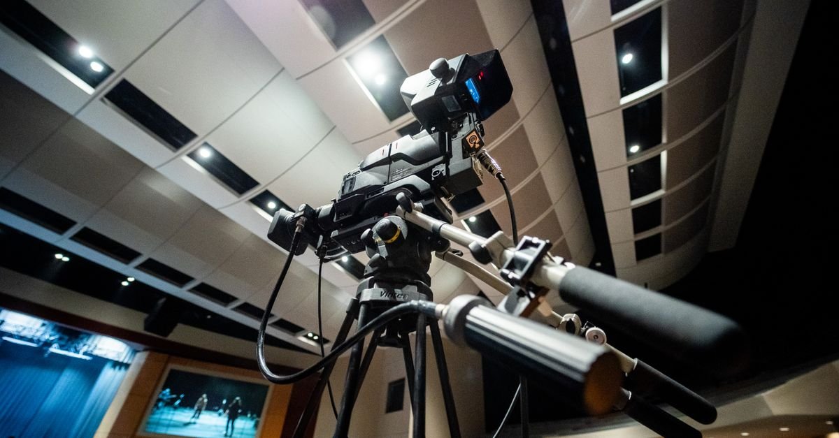 Camera de televisao sobre tripe posicionada diante de um palco de auditorio