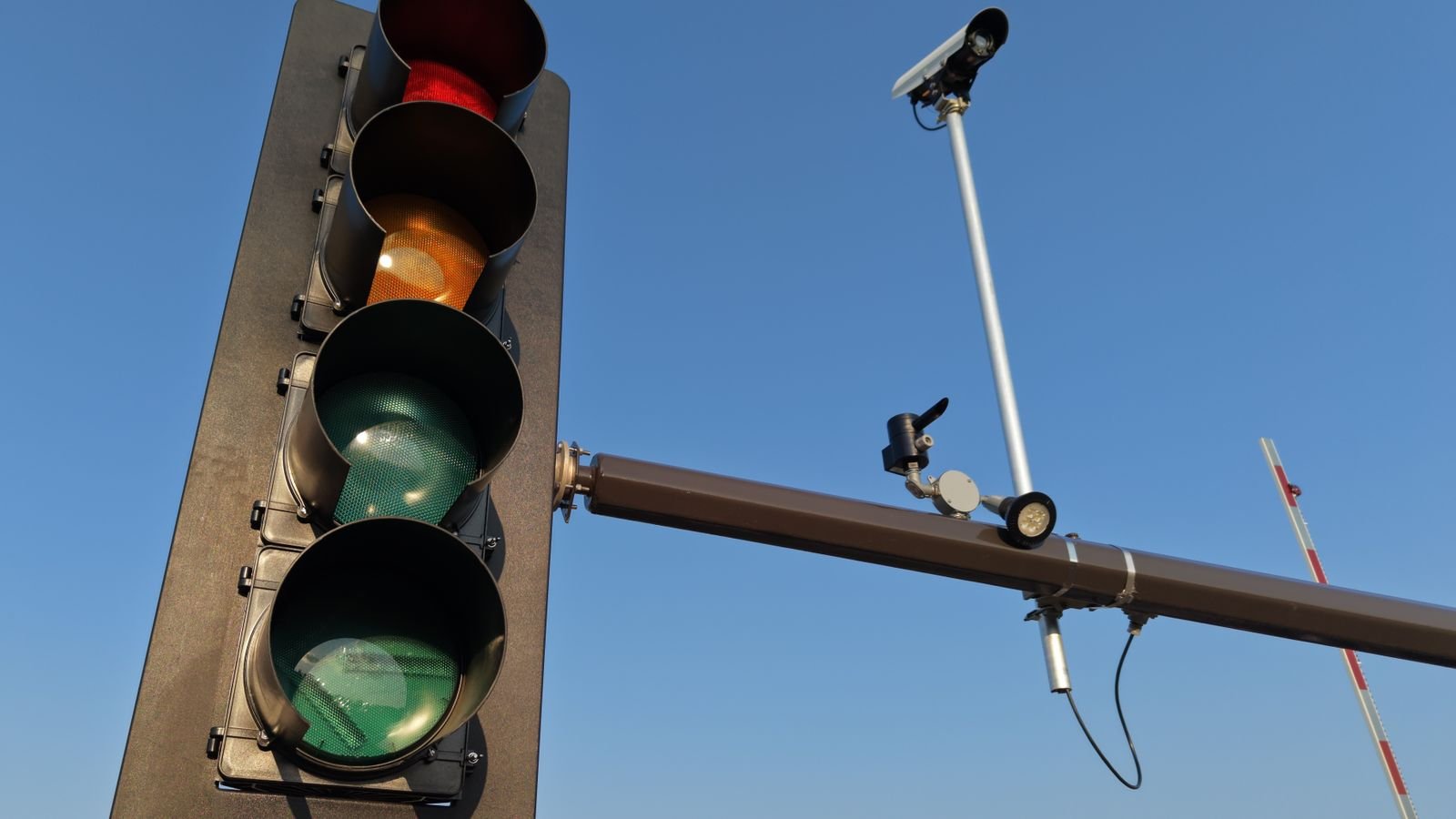 Semáforo e câmera de fiscalização de trânsito instalados no mesmo poste, vistos de baixo contra o céu azul