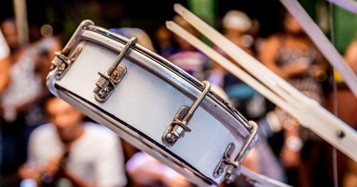 Instrumento de percussao de samba em primeiro plano durante festa ao ar livre, com publico ao fundo