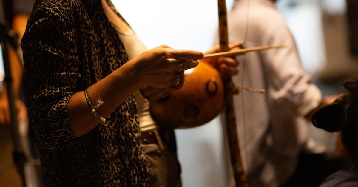 Mãos tocando berimbau, com a cabaça e a vareta em primeiro plano durante roda de capoeira