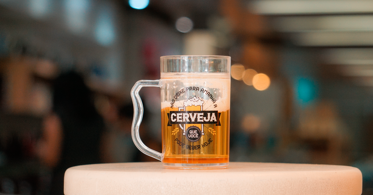 Caneca de vidro cheia de cerveja com colarinho de espuma sobre mesa, em ambiente interno de bar