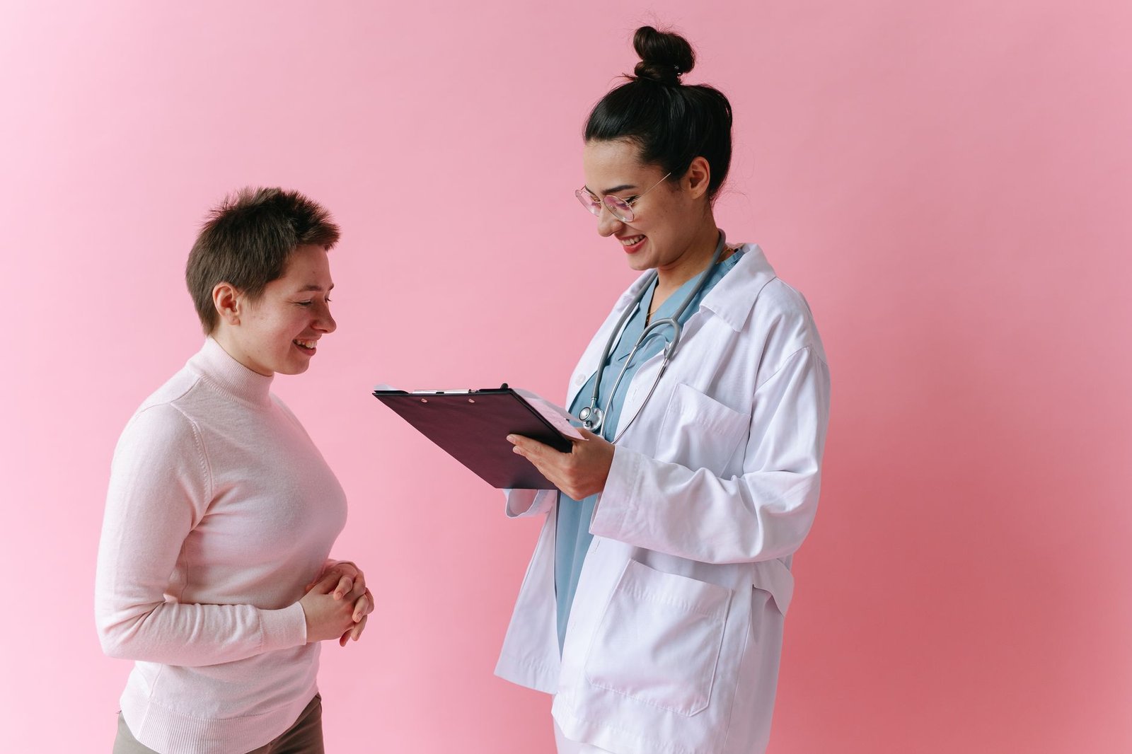 Médica sorri ao conversar com paciente em consulta, diante de fundo rosa
