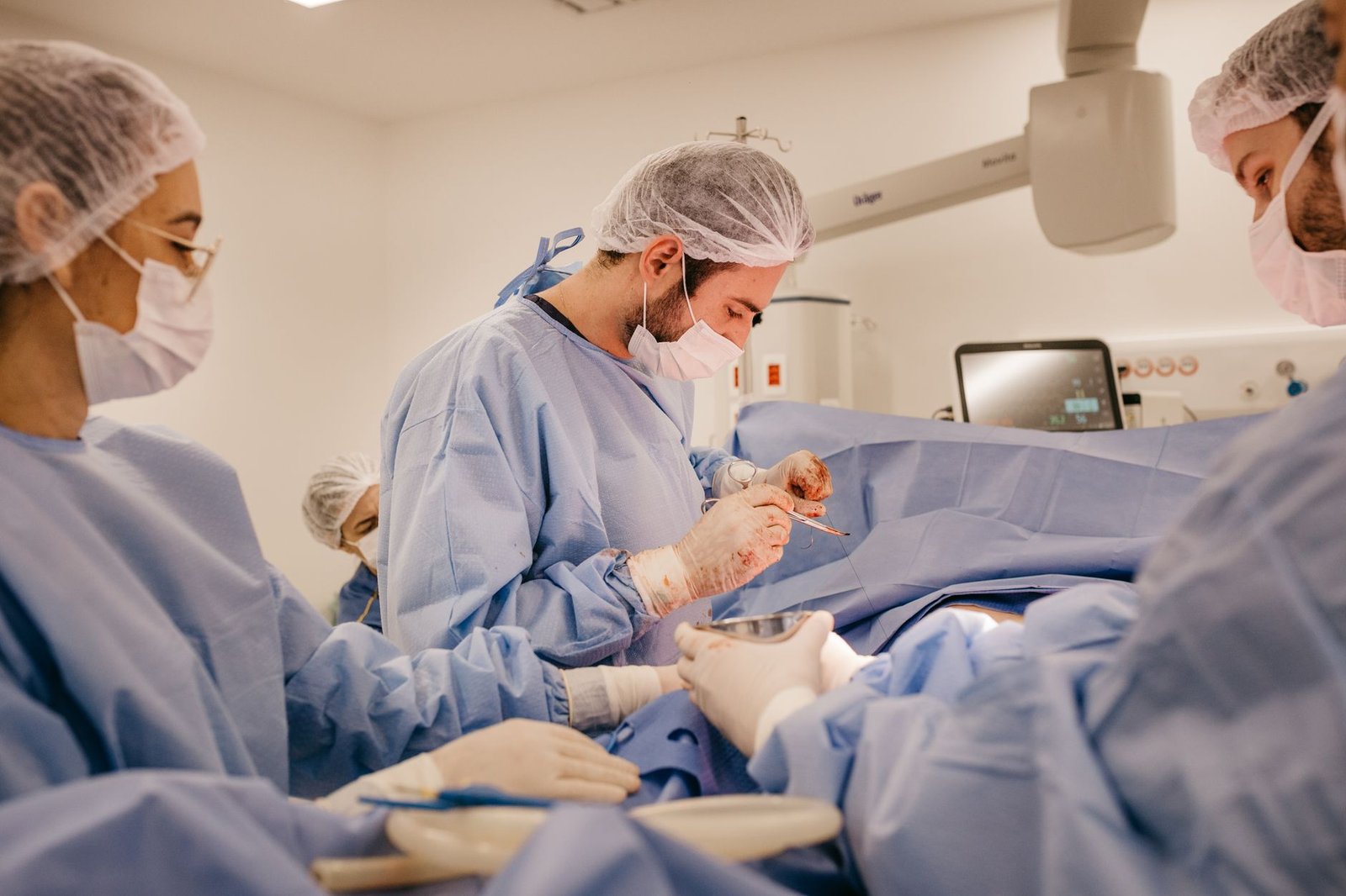 Equipe de cirurgiões com aventais azuis e máscaras durante procedimento em sala de operação, com monitor de sinais vitais ao fundo