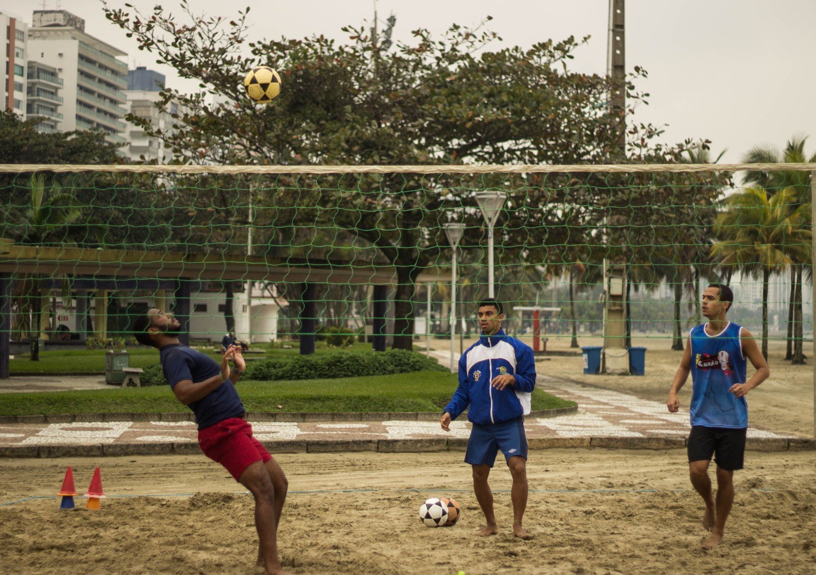 Três homens treinam futevôlei em quadra de areia ao lado de uma rede, com a bola no ar. Foto ilustrativa de uma praia brasileira, não do Oscarito.
