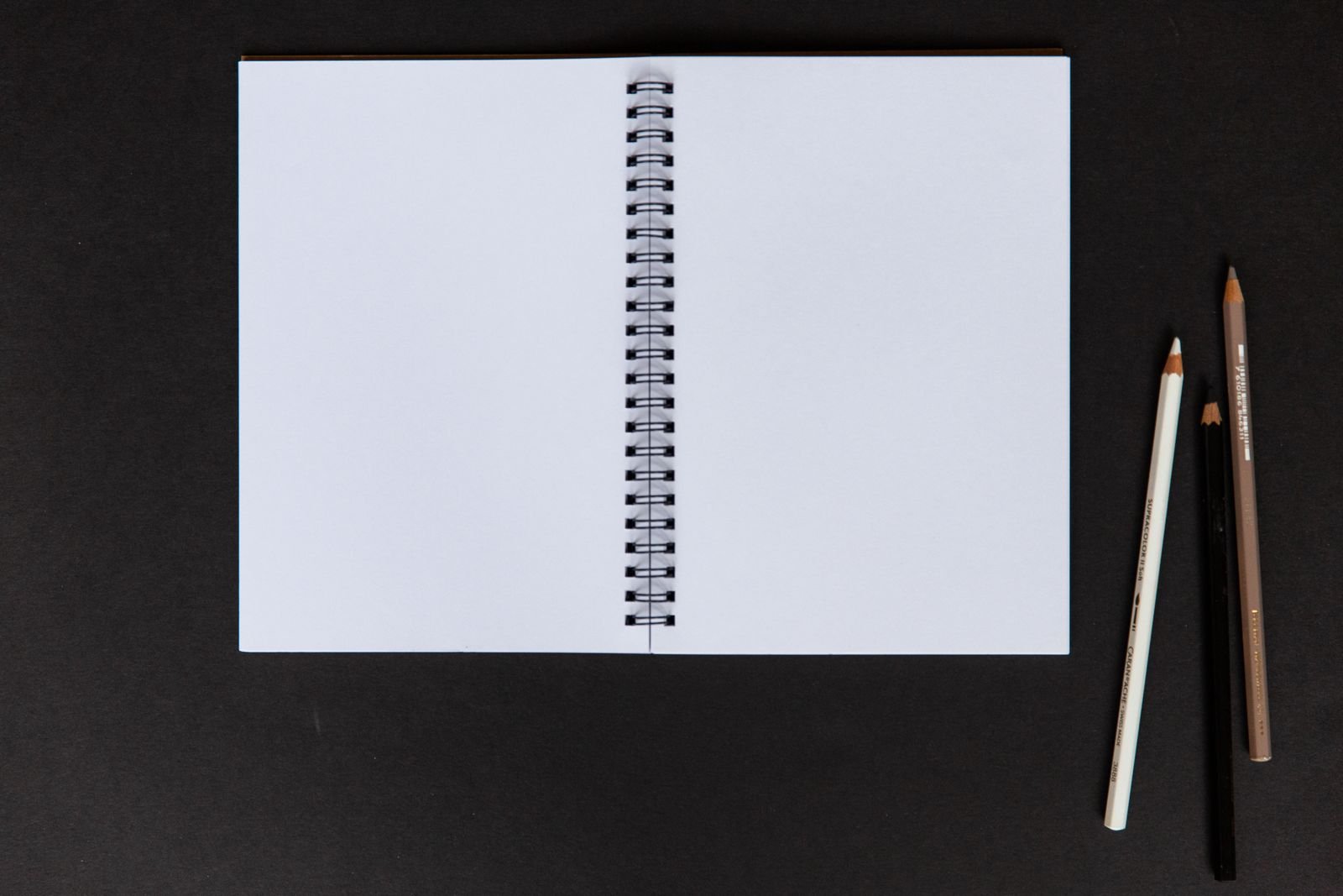 Caderno em branco aberto ao lado de três lápis sobre fundo preto
