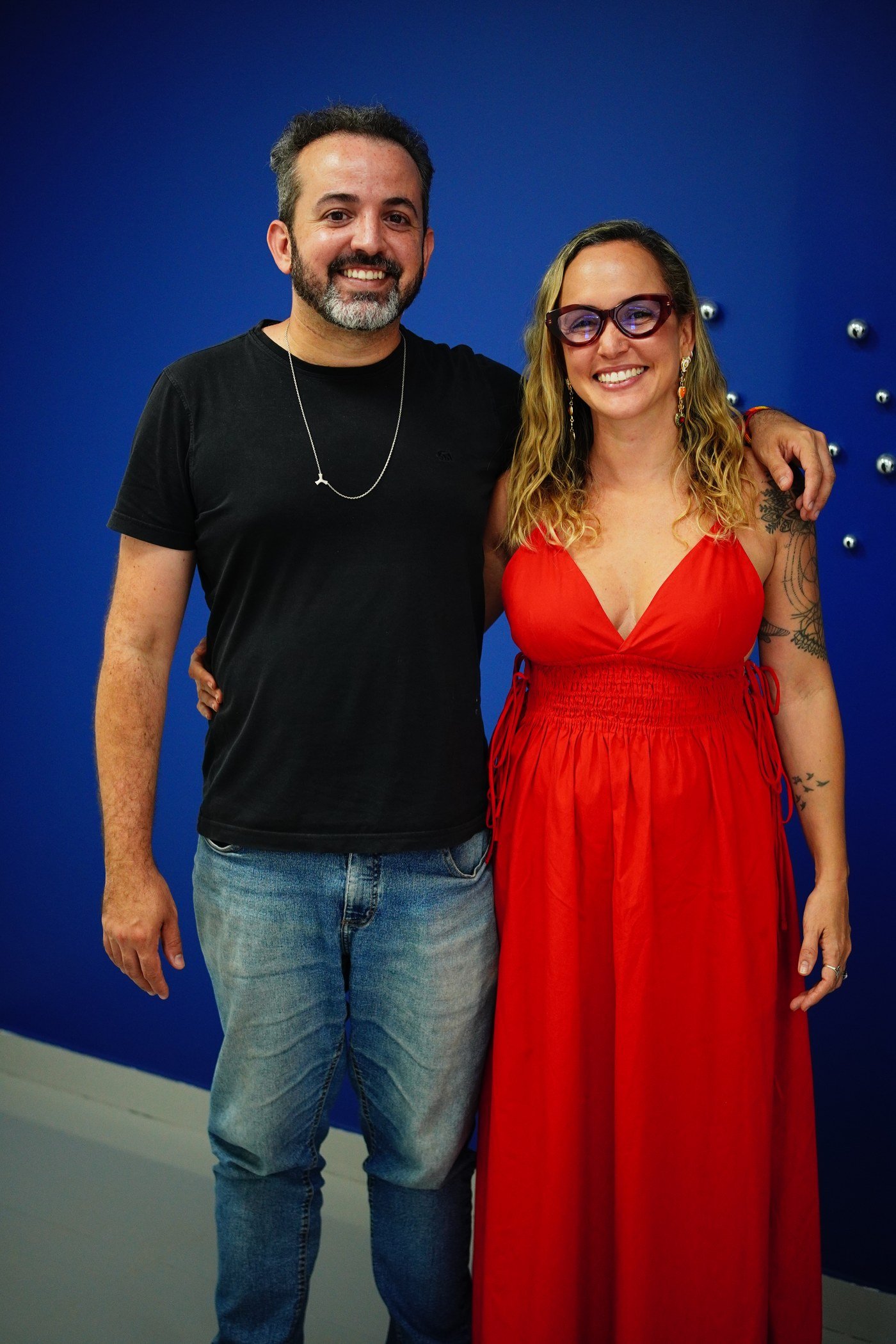 Um homem de camiseta preta e uma mulher de vestido vermelho posam abraçados diante de uma parede azul