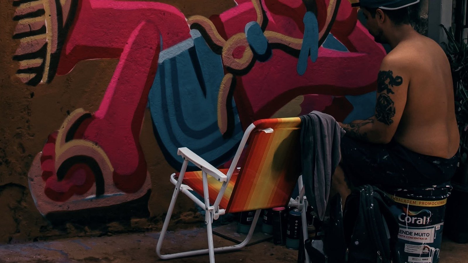 Grafiteiro de boné pinta mural colorido com figura de chapéu de palha, ao lado de cadeira de praia, latas de spray e lata de tinta