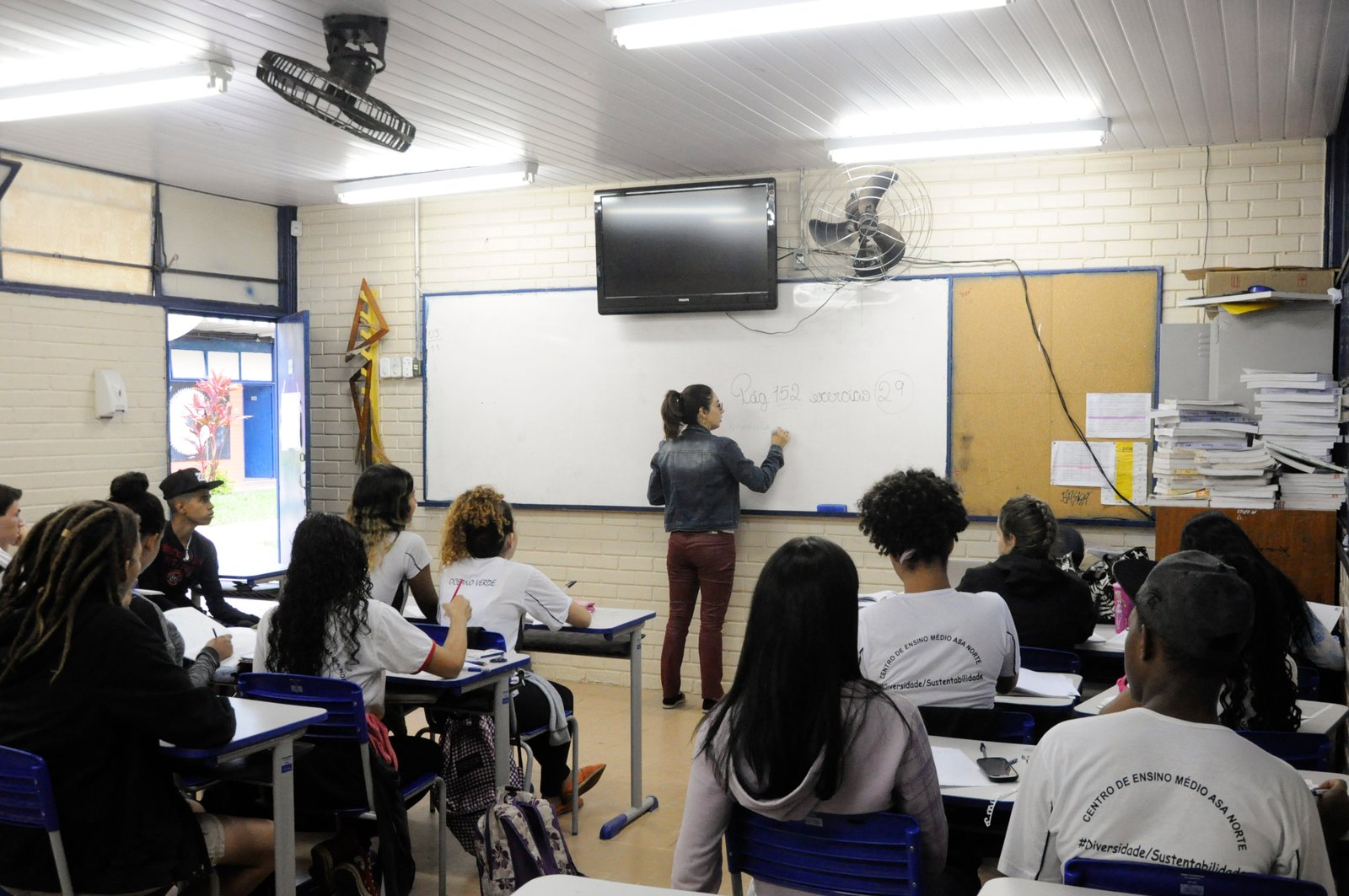 Alunos de costas em sala de aula da rede pública do DF, com professora escrevendo no quadro branco