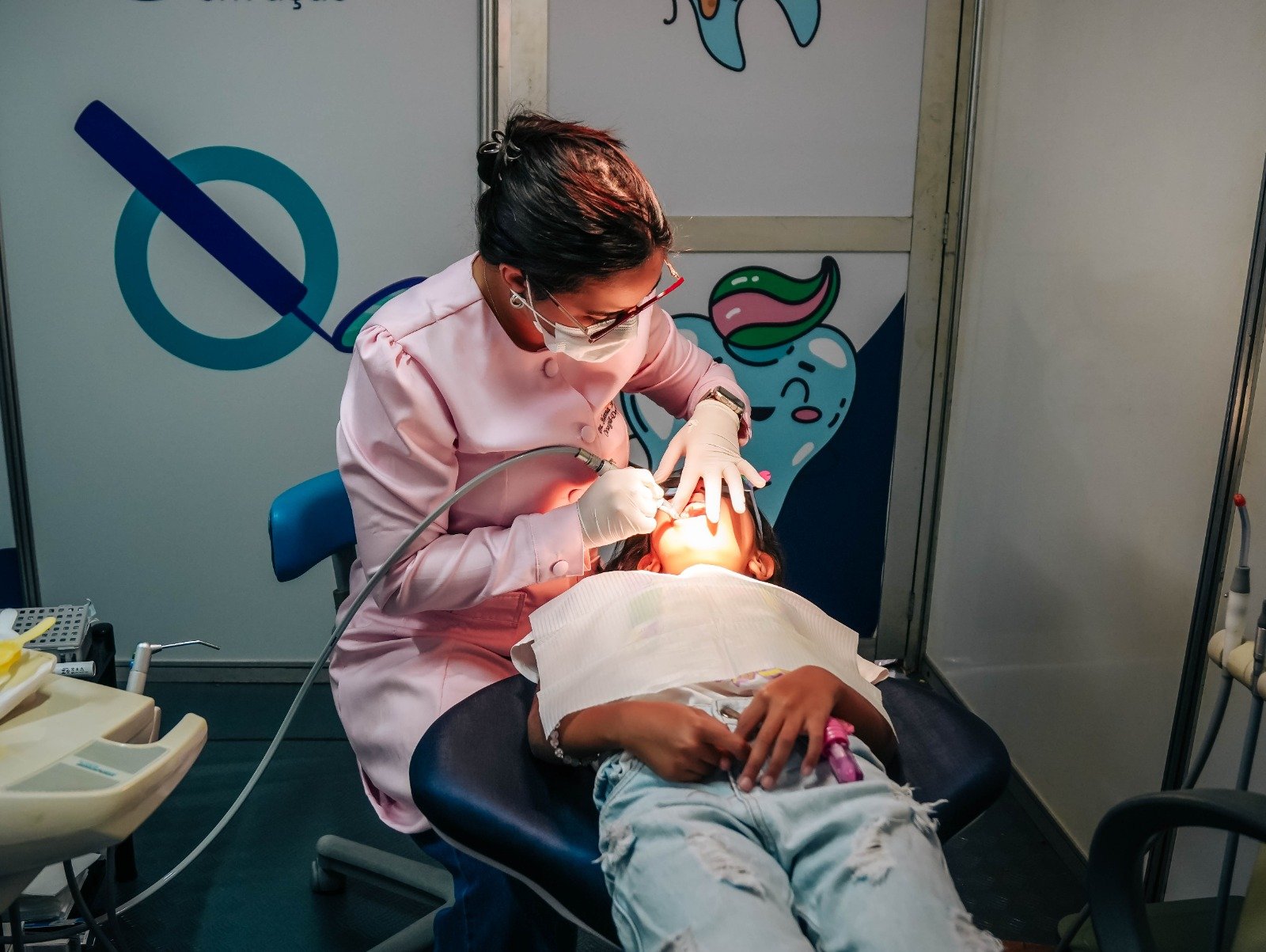 Dentista de jaleco rosa conversa com uma menina na cadeira do consultório