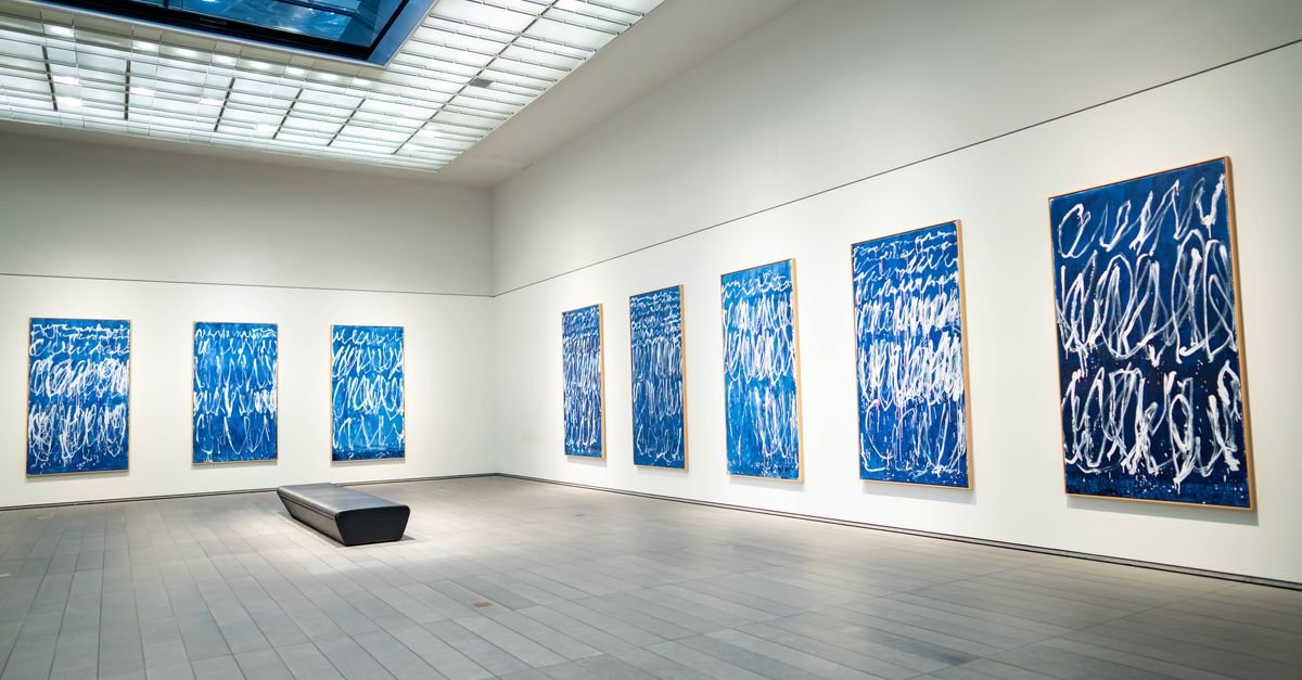 Sala de galeria de arte com telas emolduradas alinhadas nas paredes brancas e banco central
